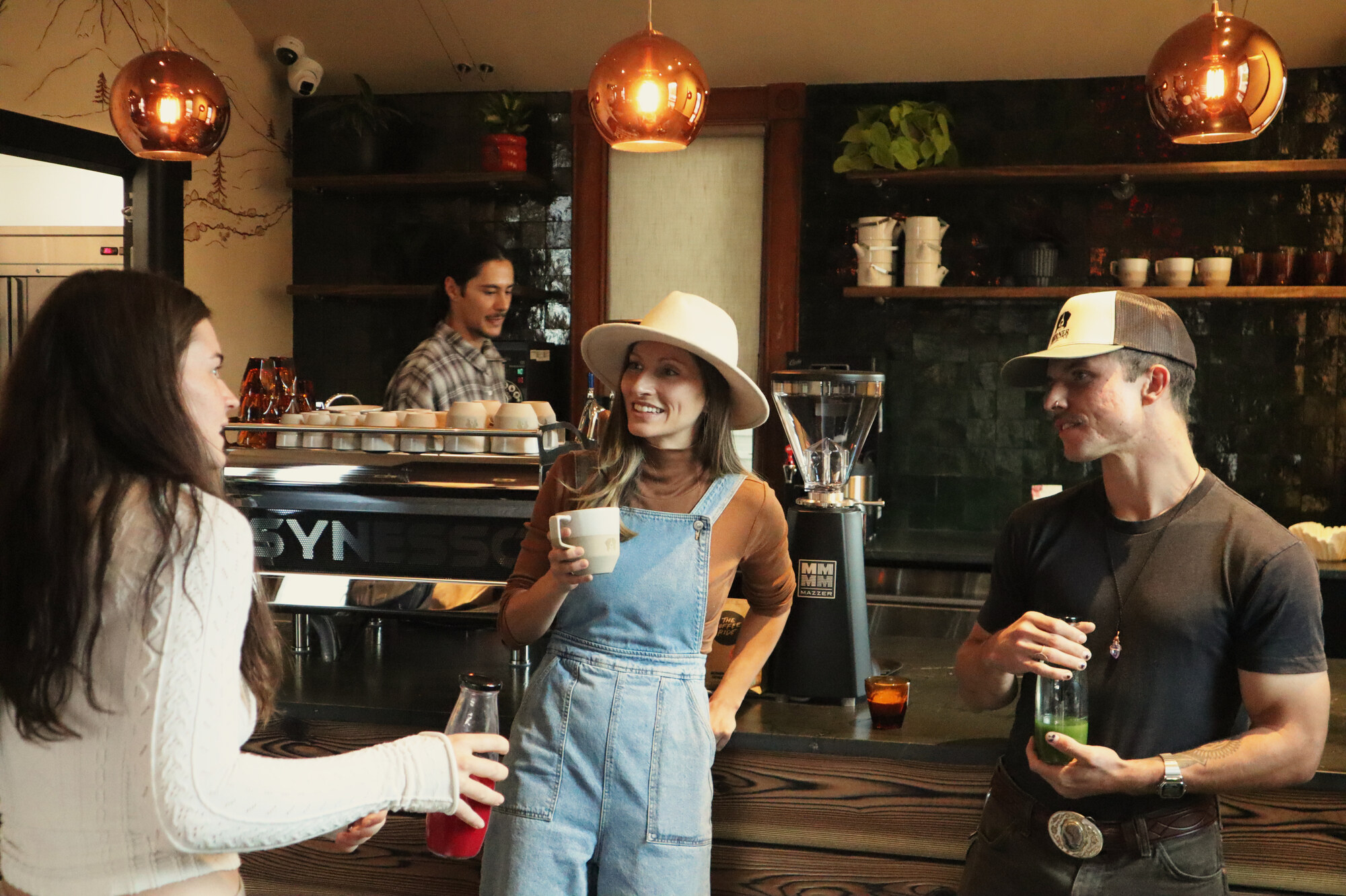 A woman in overalls and a wide-brim hat at a coffee bar